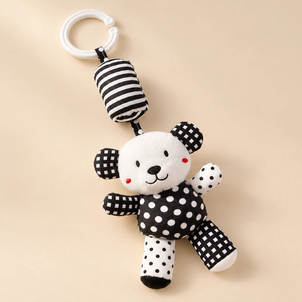 Black and white bear hanging rattle with its clip and patterned cushion on a sunlit beige tabletop.