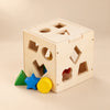 Wooden ShapeDrop sorting cube with a green triangle, blue star and yellow cylinder on a sunlit beige surface.
