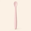 Blush silicone baby feeding spoon, showing its slim handle and shallow oval bowl on warm beige.