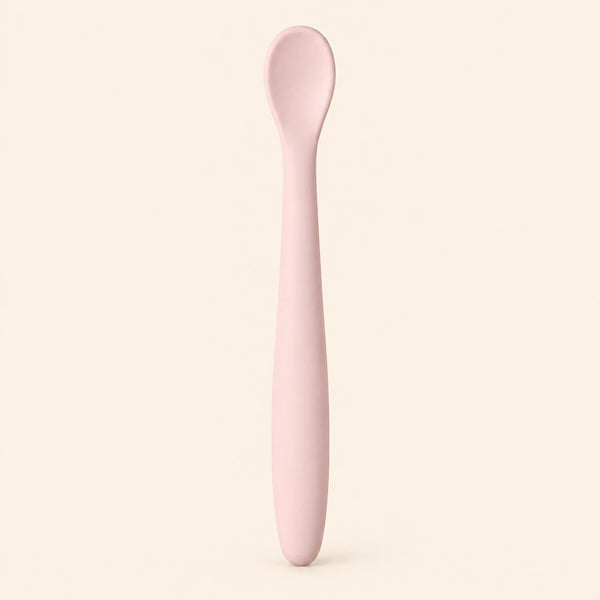 Blush silicone baby feeding spoon, showing its slim handle and shallow oval bowl on warm beige.