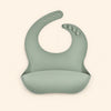 Sage Green silicone catch bib, with a scoop pocket and adjustable button-and-hole neck closure, on warm beige.