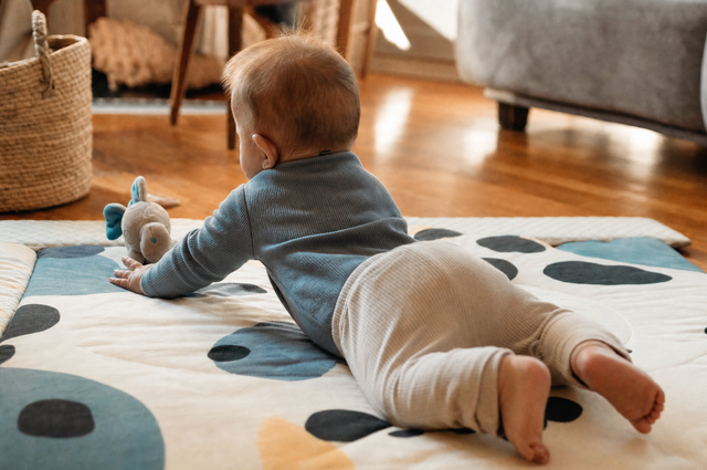 Baby on their tummy on a soft play mat, propped on forearms reaching for a toy, seen from behind