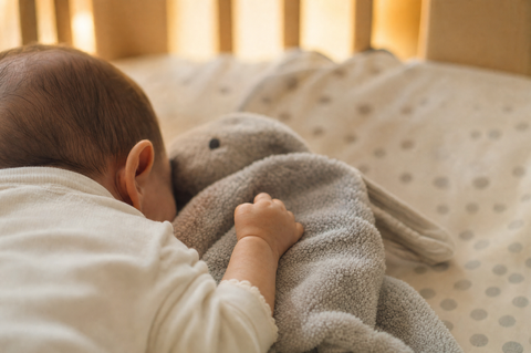 A baby's hand resting on a soft grey comfort blanket with a plush animal corner in a cozy crib