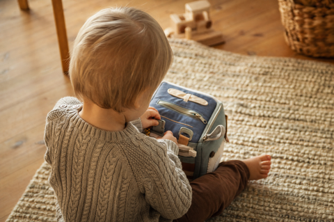 Toddler seen from behind playing alone on a rug with a soft fabric activity cube with zips and buckles