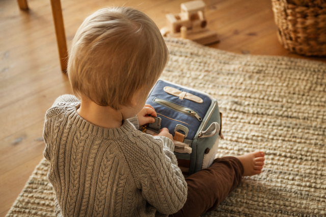 Toddler seen from behind playing alone on a rug with a soft fabric activity cube with zips and buckles