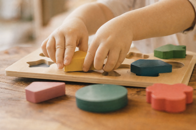 Toddler hands fitting a colorful wooden shape into a wooden shape puzzle board on a table