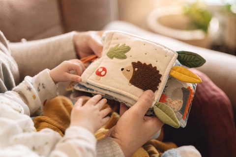 A parent's hands and a baby's hands holding a soft fabric crinkle book with animal pictures on a sofa