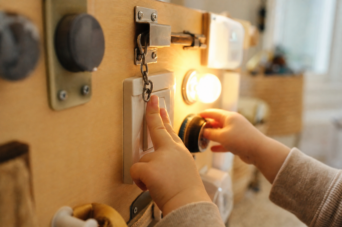 Toddler hands flipping a switch and turning a dial on a wooden busy switch board with a small light glowing
