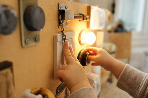 Toddler hands flipping a switch and turning a dial on a wooden busy switch board with a small light glowing