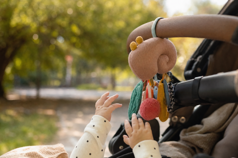 A baby's hands reaching for a colorful sensory toy clipped to a stroller bar outdoors