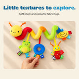 An adult hand feels a patterned fabric tag on the blue plush ring while BusyBuggy rests on a beige tabletop.