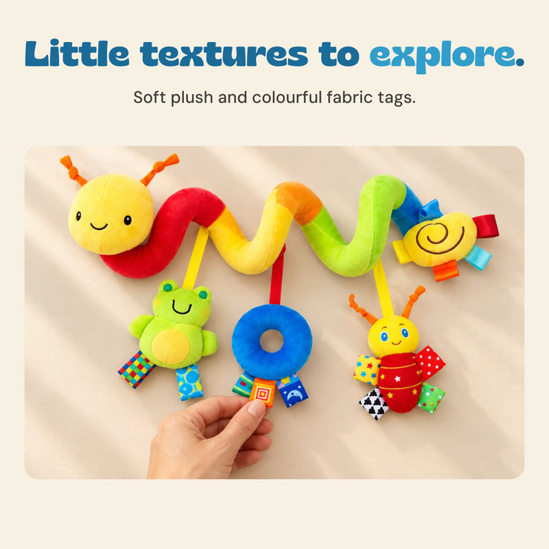 An adult hand feels a patterned fabric tag on the blue plush ring while BusyBuggy rests on a beige tabletop.