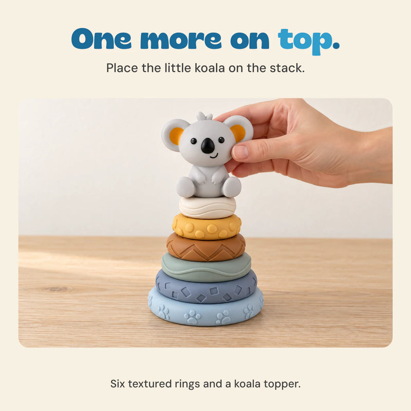 An adult hand places the grey koala topper on six textured rings on a pale wooden table.