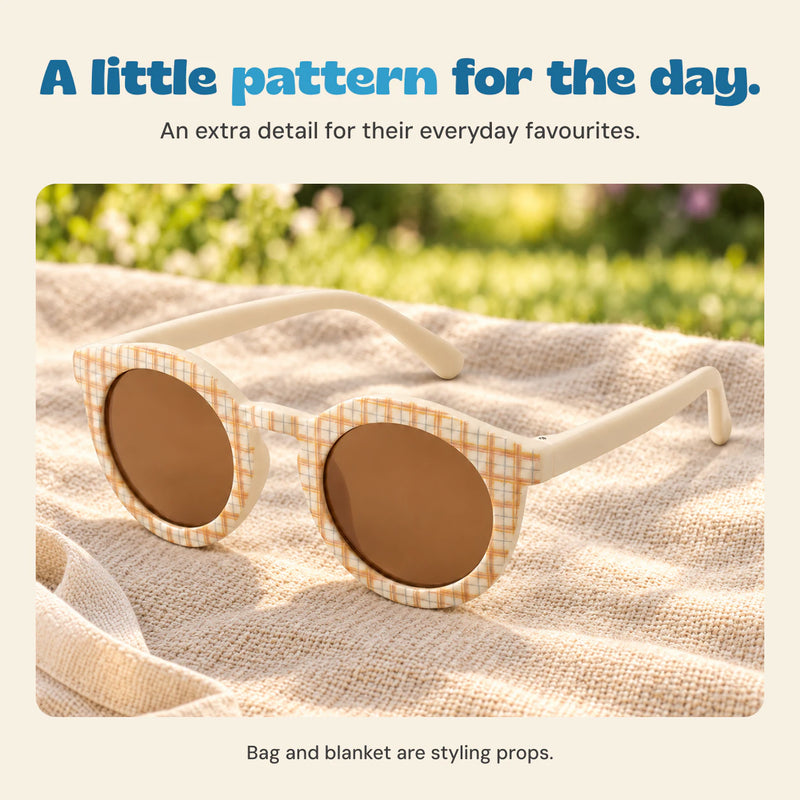 Cream plaid sunglasses resting on a beige blanket beside a canvas bag. Bag and blanket are styling props.