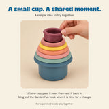 Adult hand lifting the smallest burgundy Earth Tone cup from the nested set, with a simple shared-play idea.