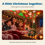 A mother and two children enjoy the Christmas tree projector in a cosy room, with the glowing tree and ring base on a bedside table.