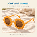 Orange flower-shaped sunglasses rest on a cream blanket beside a canvas bag. Bag and blanket are styling props, not included.