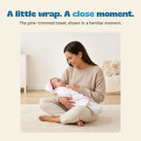 A seated mother holds an awake baby wrapped in the white towel with pink edging.