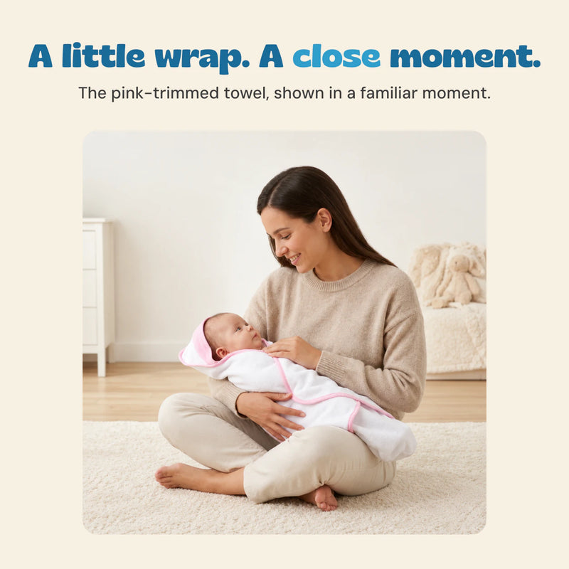 A seated mother holds an awake baby wrapped in the white towel with pink edging.