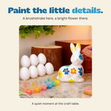 Original hand painting a blue detail on the ceramic bunny, with plain eggs and small paint pots beside it.