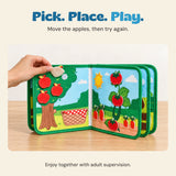 An adult hand lifts an apple piece from the Farm Friends Busy Board on a pale oak table.