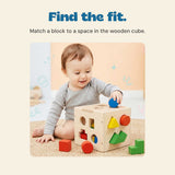 The original child places a blue round block into the wooden sorting cube, with other shapes nearby.