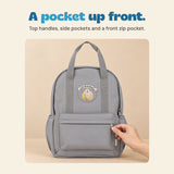 A hand holds the front-pocket zipper on the grey backpack, showing its top handles, side pockets and bear patch.