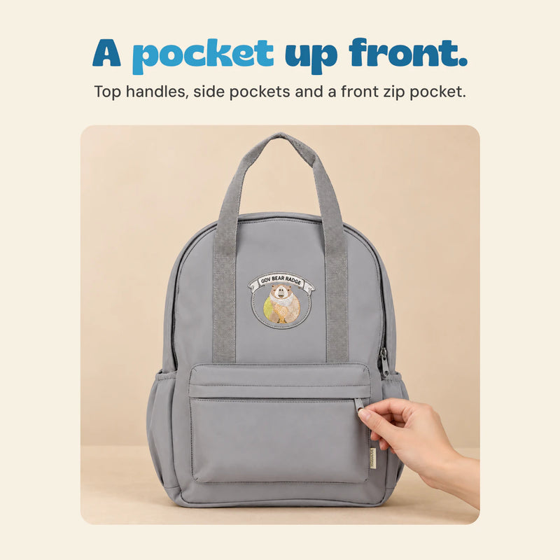 A hand holds the front-pocket zipper on the grey backpack, showing its top handles, side pockets and bear patch.