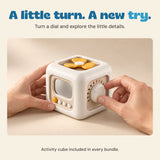 Adult hands turn the dial on the off-white and yellow activity cube on a pale oak table.