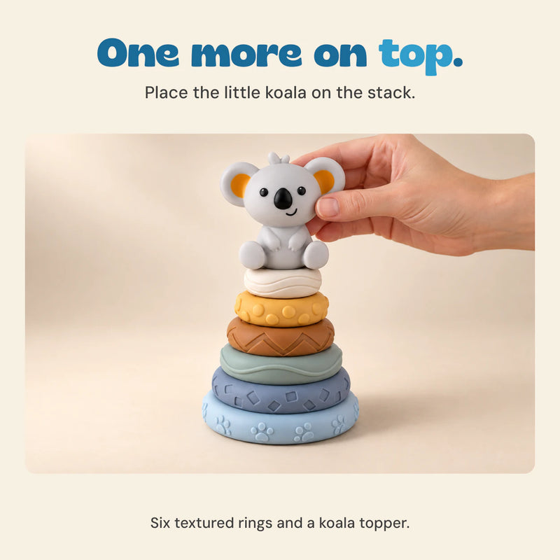 An adult hand places the grey koala topper on six textured rings on a pale wooden table.