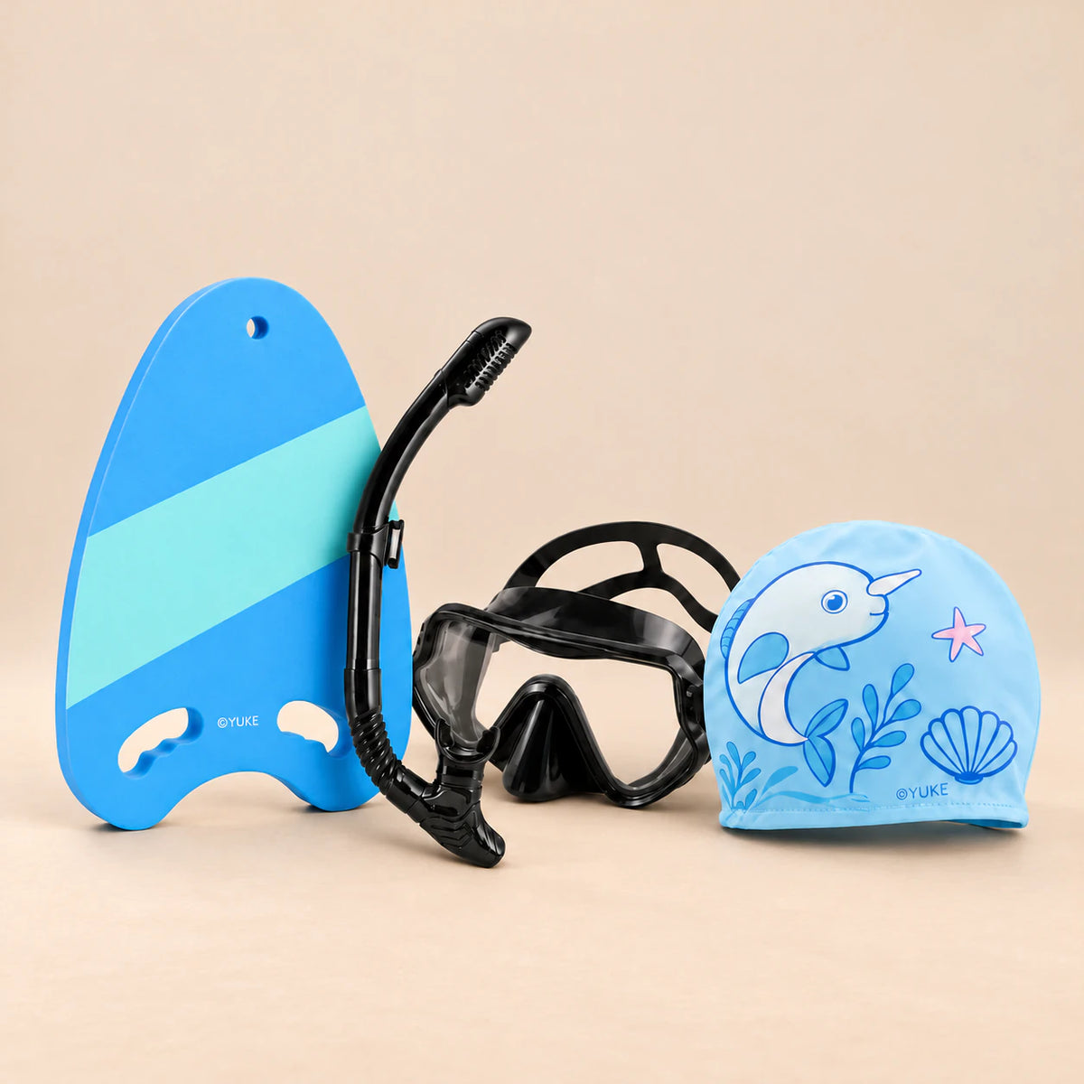 Learn-to-Swim Starter Kit with blue kickboard, black mask and snorkel, and blue narwhal swim cap on a softly lit warm beige studio surface.