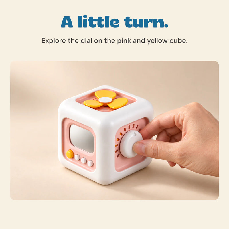 An adult hand turns the white dial on the pink and yellow Busy Cube, with mirror, buttons and yellow spinner visible.