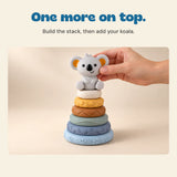 An adult hand places the grey koala on the six-ring stacking tower on a warm beige surface.