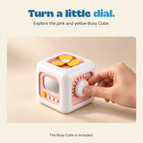 Hand turning the dial on the pink and yellow busy cube on a warm beige surface.