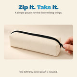 A hand holds the black zipper pull of the pale grey fabric pencil pouch on a wooden desk.