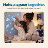 An adult and child press the glowing astronaut sticker onto a bedroom wall decorated with blue space stickers.