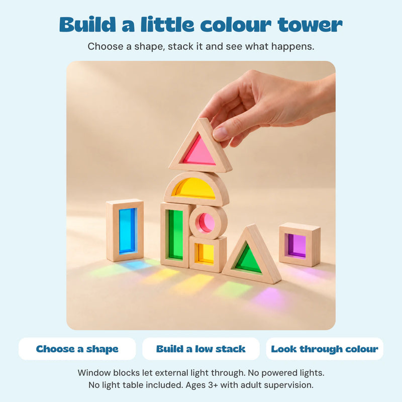 One hand stacks the wooden rainbow window blocks. Their coloured panels transmit external light; no powered lights or light table are shown.