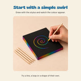 A hand using a wooden stylus to scratch a rainbow swirl on a black sheet, with four remaining wooden styluses beside the pack.