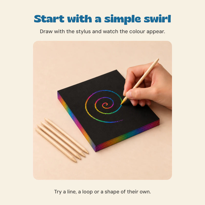 A hand using a wooden stylus to scratch a rainbow swirl on a black sheet, with four remaining wooden styluses beside the pack.