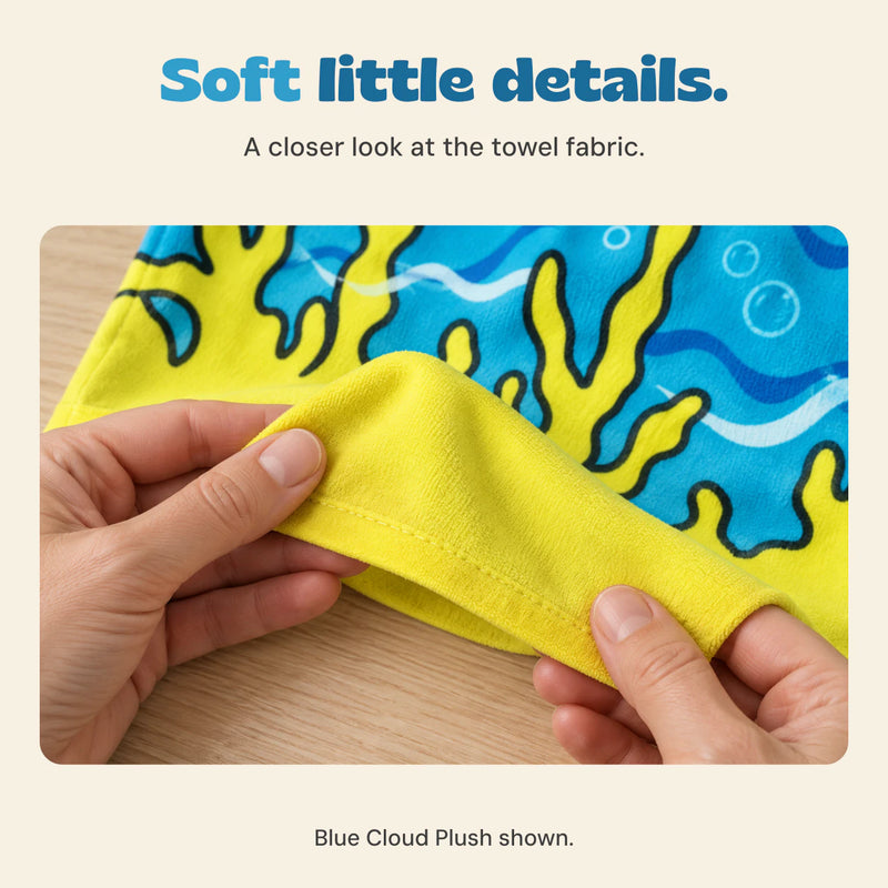 Adult hands gently hold the yellow hem of the blue shark-print microfiber poncho, showing its sewn edge and fabric.