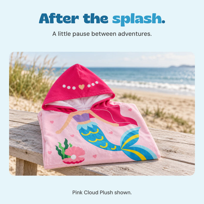 The pink mermaid poncho is folded on a pale wooden beach bench, with the printed pink hood visible.