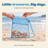 Adult hands place a shell into the blue mesh beach bag beside the sea. Shells shown for styling, bag only.