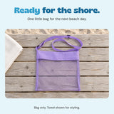 Empty purple mesh beach bag lying flat on a weathered beach bench beside an ivory towel. Towel shown for styling.