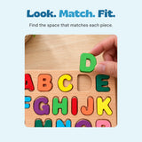 An adult hand holding the green letter D above its matching space in the wooden alphabet puzzle.