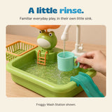 Adult hand holding a mint cup under the Froggy Wash Station tap, with the orange umbrella and turquoise slide visible.