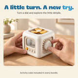 Adult hands turn the dial on the off-white and yellow activity cube on a pale oak table.