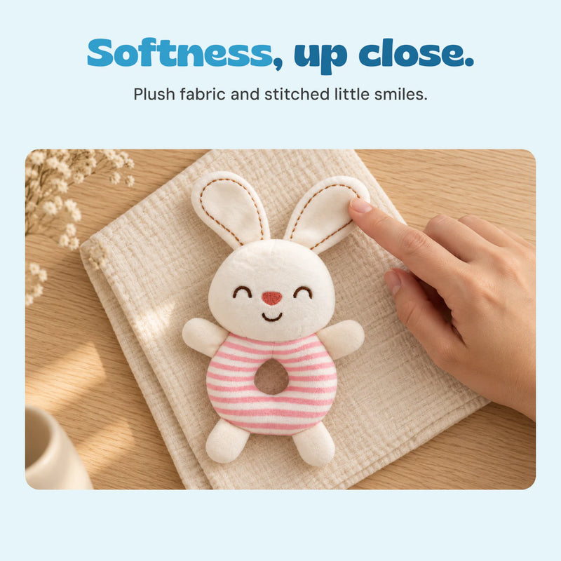 A hand touches the bunny rattle ear, showing its soft plush fabric and stitched details.