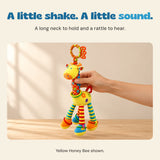 An adult hand holds the yellow giraffe rattle by its long neck above a wooden table.