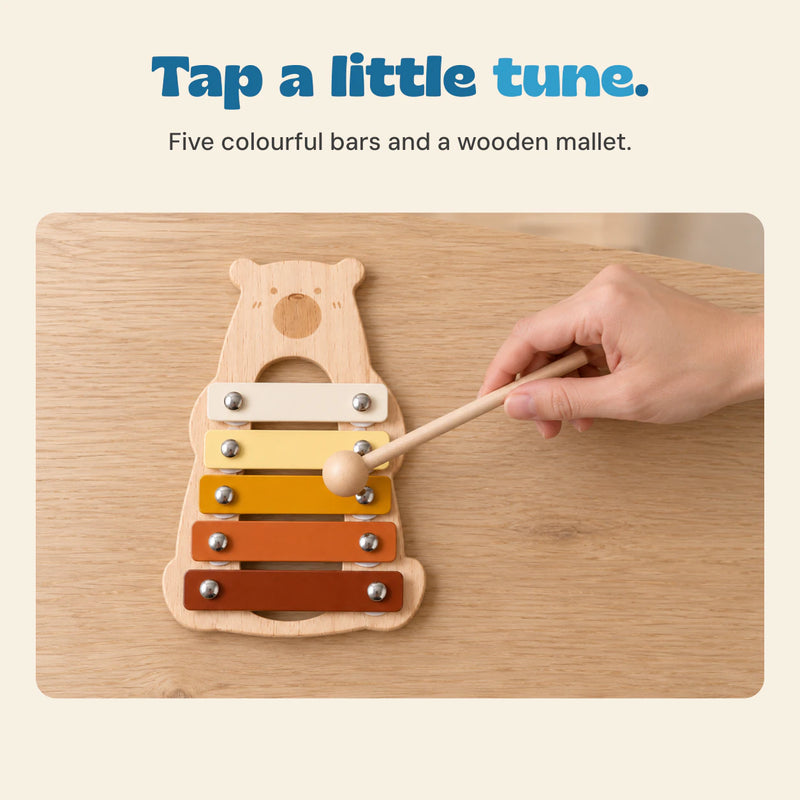 An adult hand holds the single wooden mallet above the five metal bars of the bear-shaped xylophone lying flat on an oak table.