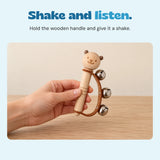 Adult hand holding the wooden bear bell rattle, with its curved brown strap and three silver bells visible.